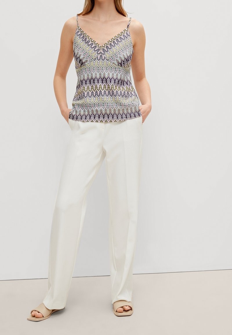 Mönstrad ärmlös linne i nyanser av lila, grönt och krämvitt, kombinerad med lösa vita byxor och beiga slip-in sandaler.