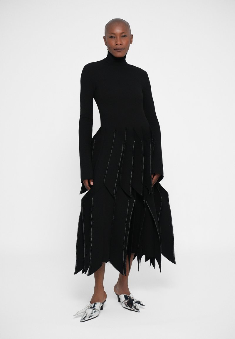 Black long-sleeve dress featuring a fitted bodice with layered, jagged hems and contrasting stitching. Paired with silver heeled mules.
