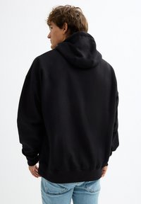 Sudadera con capucha negra con bolsillo canguro, corte oversize y puños acanalados. Tejido suave con textura lisa, vista desde atrás.