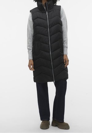 Personne portant une longue veste matelassée noire sur un pull gris, un pantalon foncé et des bottes vert olive, debout devant un fond uni.