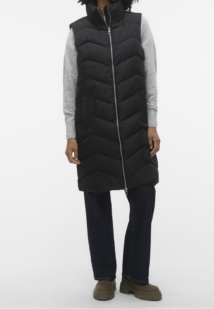 Personne portant une longue veste matelassée noire sur un pull gris, un pantalon foncé et des bottes vert olive, debout devant un fond uni.