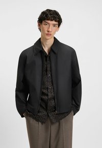 Black lightweight jacket with a collared neckline, zip closure, and relaxed fit. Features a patterned shirt underneath and neutral-toned trousers.