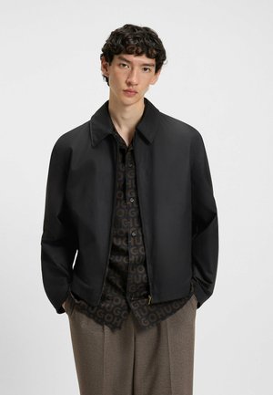 Giacca nera leggera con collo a camicia, chiusura con zip e vestibilità morbida. Presenta una camicia a fantasia sotto e pantaloni in tonalità neutra.