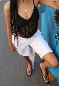 Zwarte kanten bodysuit met een gegolfde halslijn, gecombineerd met witte denim shorts. Draagt zwarte slippers en staat naast een blauwe auto.