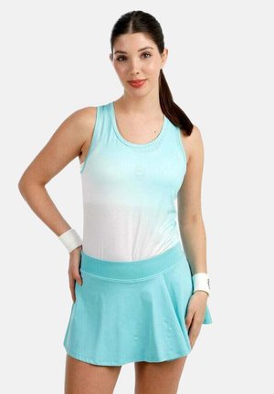 Jonge vrouw met een donkere paardenstaart, gekleed in een lichtblauw mouwloos topje, bijpassende rok en witte polsbandjes, staande met een hand op haar heup.