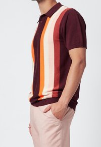 Mies, joka on pukeutunut lyhythihaiseen poolopaitaan, jossa on burgundin, oranssin, vaaleanpunaisen ja kermanväriset pystysuorat raidat, sekä vaaleanpunaisiin housuihin, ja hänellä on käsi taskussa.