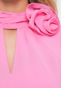 Abito rosa con leggero scollo a V e dettaglio di tessuto arricciato. Presenta un grande accento di rosa stratificato sulla spalla sinistra. Texture morbida e liscia.