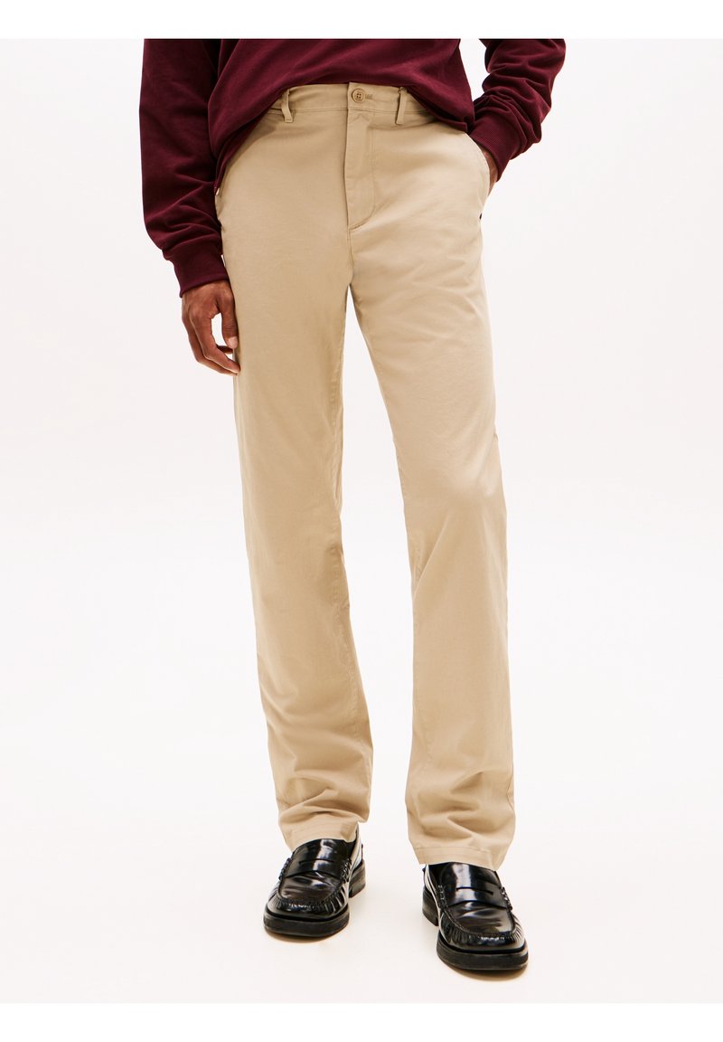 Homme portant un pantalon beige droit, des mocassins en cuir noirs et une chemise à manches longues rouge foncé, debout avec une main dans la poche.