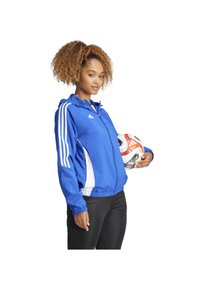 Blauwe sportjack met witte zijkanten, volledige rits en capuchon. Heeft drie witte strepen op elke mouw. Houdt een voetbalsbal vast.