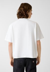 Biała koszulka z krótkim rękawem o luźnym kroju, gładkiej strukturze i prążkowanej tkaninie. Posiada okrągły dekolt oraz małą naszywkę z logo przy dolnym brzegu.