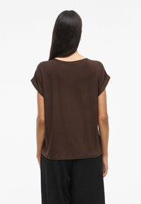 Brūns īsām piedurknēm tops ar brīvu piegriezumu, noapaļotu kakla izgriezumu un sarullētām piedurknēm, izgatavots no mīksta auduma. Aizmugures skats parāda gludu tekstūru un skaistu krāsu.