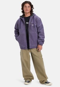 Purple hooded jacket made of denim, with front pockets and elastic cuffs. Paired with loose beige pants and black sneakers.