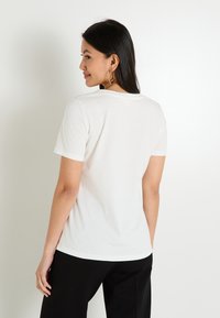 T-shirt en coton blanc à manches courtes et col rond, avec un ourlet arrondi et une texture lisse. Porté avec un pantalon noir.