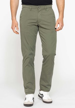 Uomo che indossa pantaloni casual verde oliva e sneakers bianche con strisce nere, in piedi davanti a uno sfondo chiaro uniforme.