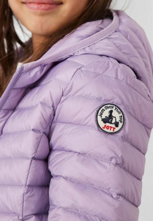 Fille portant une veste matelassée violet clair avec capuche, montrant un patch rond sur la manche avec une silhouette de trottinette et le texte « Just Over The Top JOTT ».