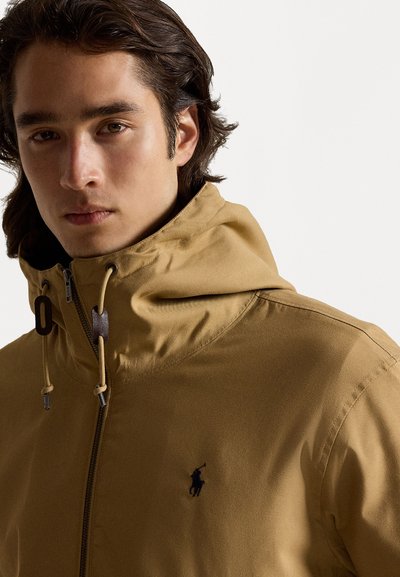 Tan hooded jacket with a front zipper, drawstring accents, and a small black logo on the left chest. Smooth, matte texture.