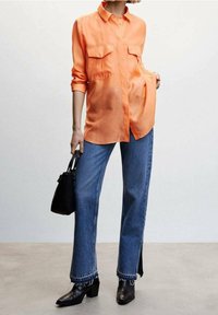 Camicia arancione a maniche lunghe con due tasche sul petto, abbinata a jeans blu a vita alta e gamba larga, e stivaletti neri con tacco. Borsa a mano nera come accessorio.
