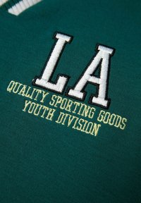 Grøn stof med hvid og gul broderet tekst: "LA QUALITY SPORTING GOODS YOUTH DIVISION," med et fremtrædende logodesign.