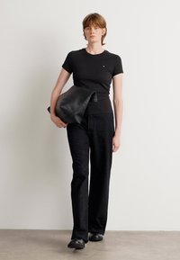 T-shirt noir ajusté avec un petit logo, associé à un pantalon large noir légèrement scintillant. Elle porte un sac en cuir noir.