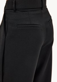 Pantalones negros de corte entallado que presentan una textura de tela suave, diseño ajustado y dos bolsillos traseros. La cinturilla es plana, realzando la silueta.