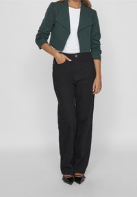 Woman wearing a dark green cropped blazer, white top, high-waisted black pants, and black pointed-toe heels, standing with one hand in pocket.