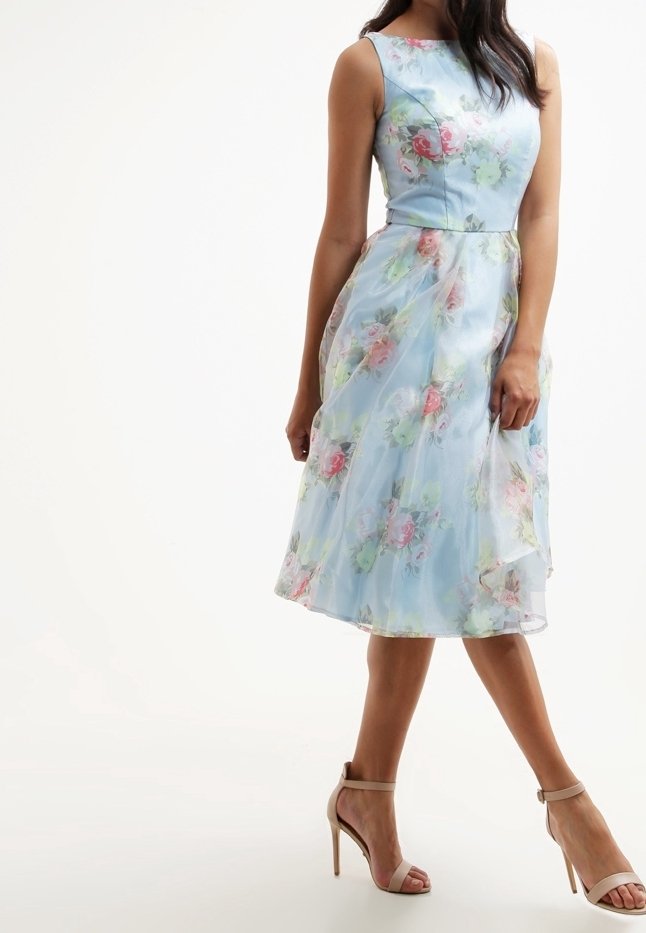 Vaaleansininen kukkakuvioinen mekko, jossa on istuva vartalo ja laskeutuva helma. Mekossa on pastellinvaaleanpunaisia ja vihreitä kukkia, hihaton malli ja läpikuultava päällyskerros.