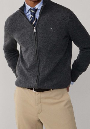 Man draagt een donkergrijze trui met rits over een blauw gestreept overhemd en een gestreepte stropdas, gecombineerd met beige broek en zwarte riem.