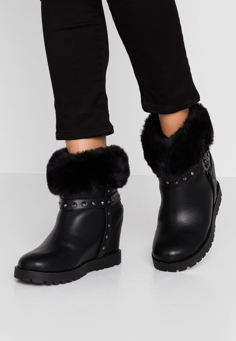 Bottines noires à talon compensé avec manchette en fourrure et bride cloutée portées avec un pantalon noir sur fond clair.