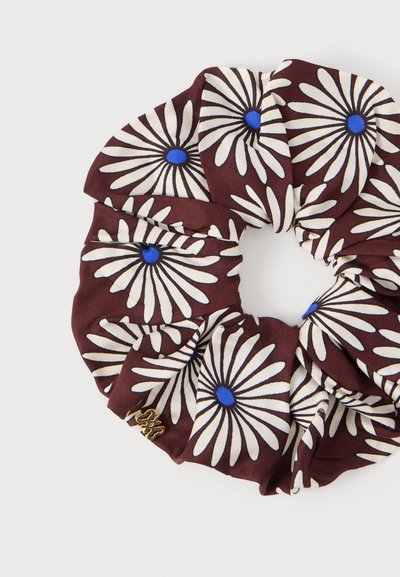 Chouchou en tissu marron avec des marguerites blanches aux centres bleus et un petit charm décoratif doré.