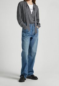 Blusa negra con estampado y mangas largas, atada en la parte delantera, combinada con jeans de tiro alto en color azul claro y zapatos negros con suela gruesa.