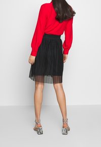 Schwarzer Faltenrock mit elastischem Bund, kombiniert mit einer roten Bluse mit langen Ärmeln und schwarz-weiß gestreiften Sandalen mit Absatz.