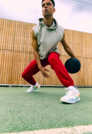 Valge sportlikud jalanõud erevärviliste aktsentidega, hall kapuutsiga pusa ja punased spordipüksid, dribblimas musta korvpalli rohelisel väljakul.