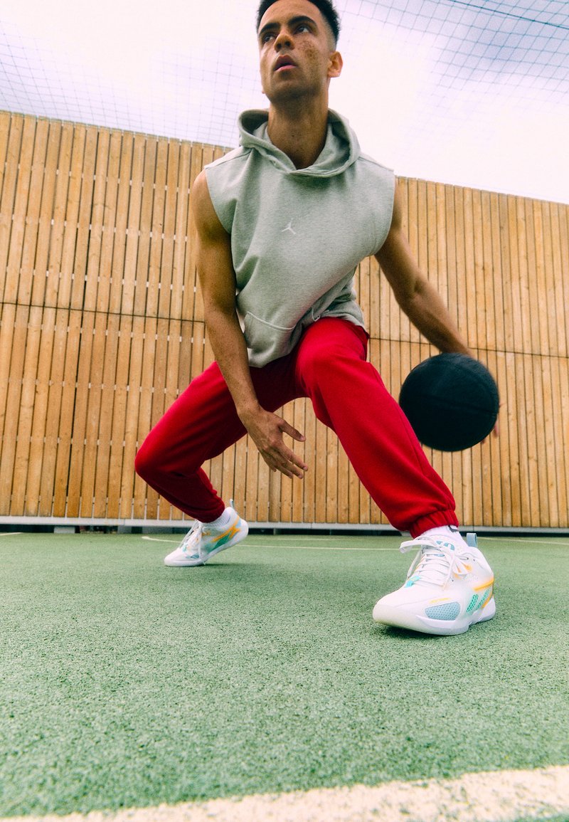 Weiße Sportschuhe mit bunten Akzenten, grauer Kapuzenpullover und rote Sweatpants, die einen schwarzen Basketball auf einem grünen Platz dribbeln.