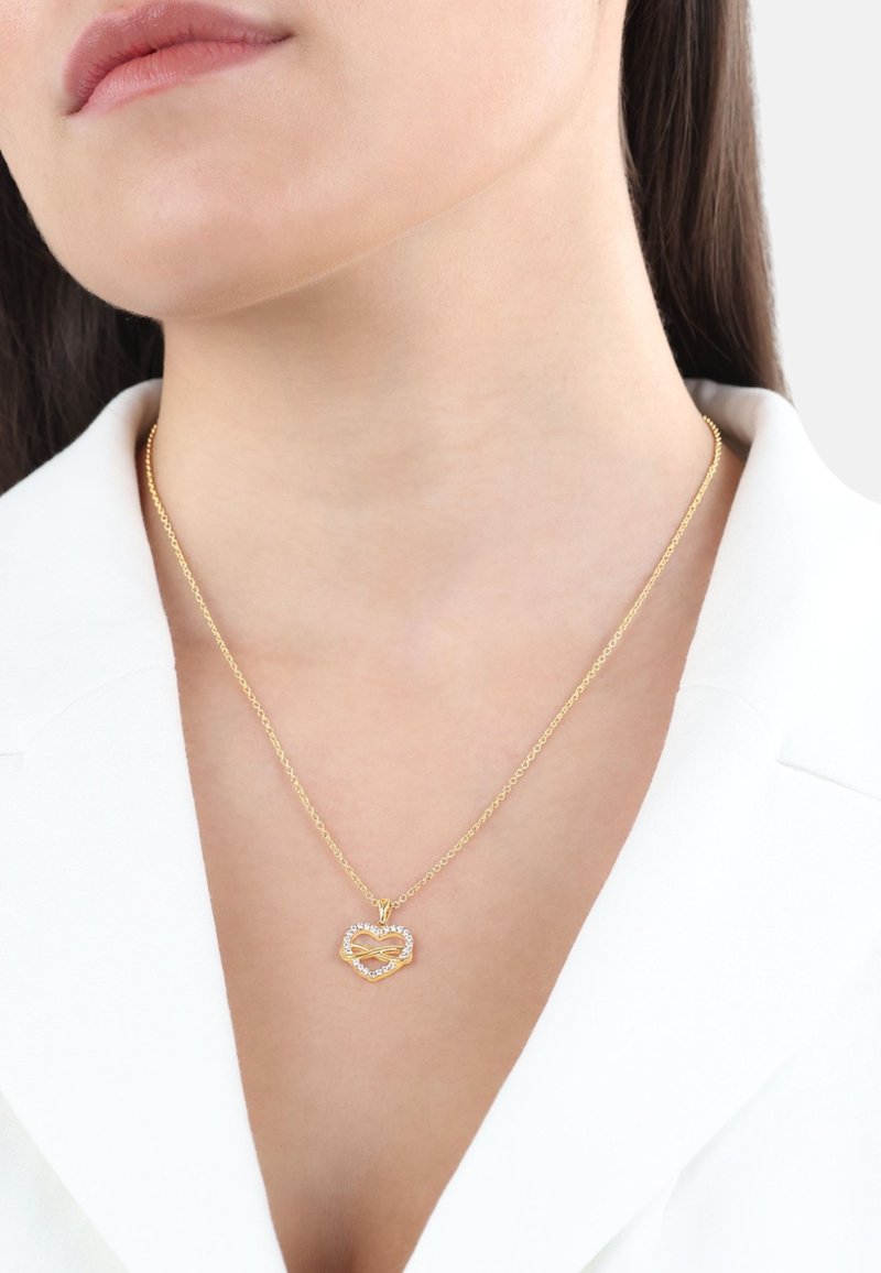 Collana in oro con ciondolo a forma di cuore e simbolo dell'infinito, arricchita da pietre trasparenti, indossata sopra un blazer bianco.