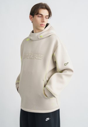 Jeune homme portant un sweat à capuche beige Nike avec des accents néon, les mains dans les poches, regardant sur le côté, assorti à un pantalon noir Nike.