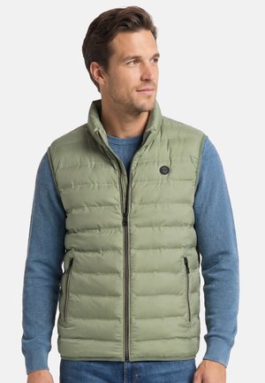 Mann trägt eine olivgrüne gesteppte Weste über einem blauen Pullover, schaut nach rechts, mit sichtbarem Reißverschluss und Reißverschlusstaschen.