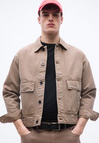 Νεαρός άνδρας με ανοιχτό καφέ σακάκι με κουμπιά, μαύρο πουκάμισο, ταιριαστό παντελόνι, μαύρη ζώνη και κόκκινο καπέλο μπέιζμπολ, με τα χέρια στις τσέπες.