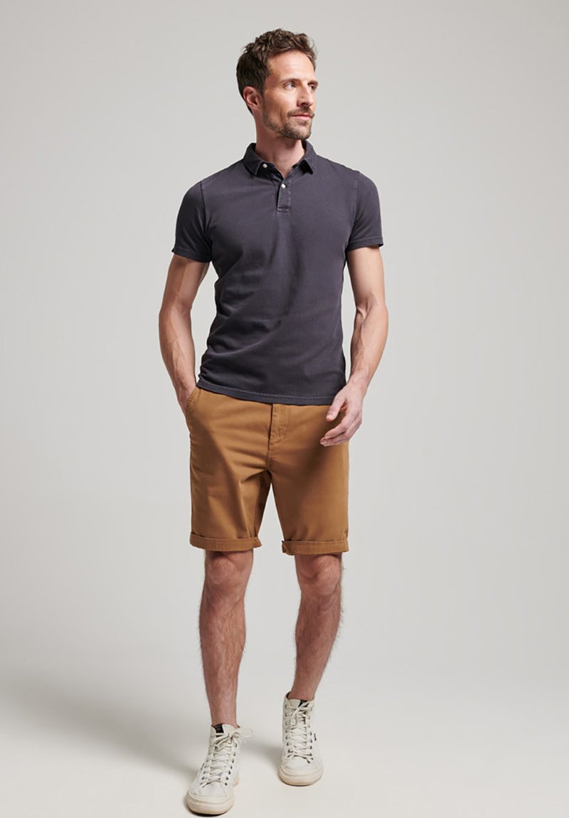 Camisa polo de manga corta en color azul marino, pantalones cortos marrones con dobladillo y zapatillas blancas. La polo tiene un cuello y un diseño ajustado; los pantalones cortos son de frente plano.