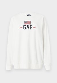 Sweatshirt blanc à manches longues, col rond, comportant un logo brodé "GAP" et un design de drapeau américain en rouge et bleu.