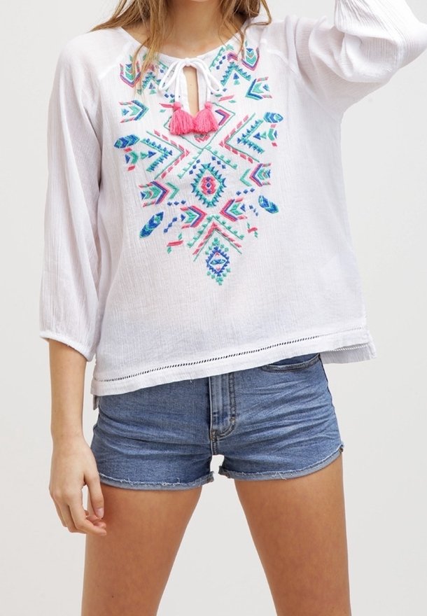 Femme portant un chemisier blanc à manches longues avec une broderie colorée et des pompons roses, associé à un short en jean bleu.