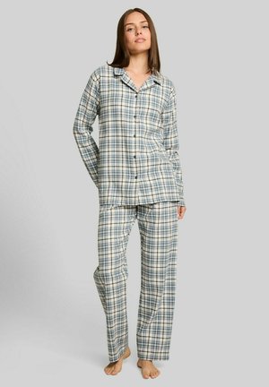 Jonge vrouw staat op blote voeten, draagt een blauw-wit geruit pyjamablouse met knopen en lange mouwen en bijpassende broek op een effen achtergrond.
