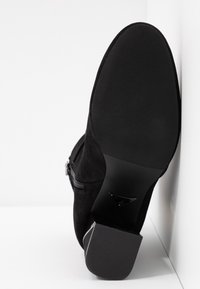 Semelle et talon d'une botte cheville en daim noir avec semelle en caoutchouc texturé et fermeture éclair latérale, présentés reposant contre une surface blanche.