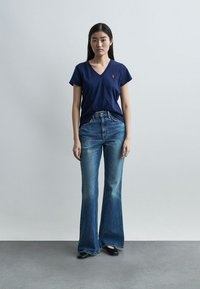 Νέα γυναίκα που στέκεται μπροστά σε απλό φόντο, φορώντας μπλε μπλούζα V-neck, μπλε ψηλόμεσα φαρδιά τζιν και μαύρα παπούτσια.