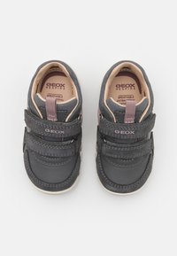 Chaussures gris foncé en matériau respirant, avec deux brides Velcro, des accents beiges et une semelle en caoutchouc robuste avec un motif texturé.