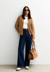 Beige stickad cardigan, vit t-shirt, mörkblå vida jeans, vita sneakers, brun mockaväska och spegelglasögon på en modell.