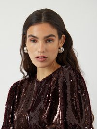 Top di un profondo borgogna in paillettes con spalle arricciate e collo alto. Orecchini a cerchio in argento accentuano il look. Texture liscia e riflettente.