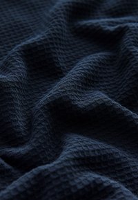 Mørkeblå stof med et tekstureret vaffelmønster vist i nærbillede med bløde folder og skygger.