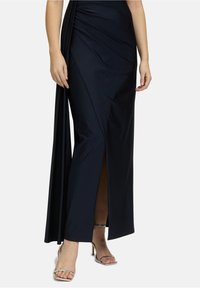 Robe noire longue jusqu'au sol avec tissu drapé et fente sur le côté. Texture lisse et design ajusté mettant en valeur des talons à lanières argentés.