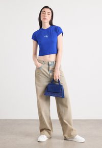 Femme aux longs cheveux noirs se tient dans une posture détendue, portant un t-shirt bleu court et un pantalon large beige, tenant un sac à main bleu.