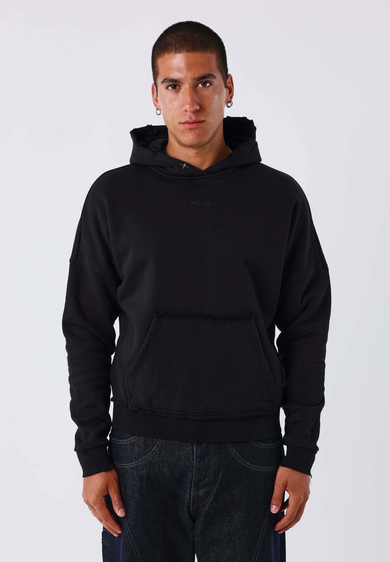 Jeune homme portant un sweat à capuche noir avec une poche avant et un jean foncé, debout devant un fond clair uni, faisant face à l'avant.
